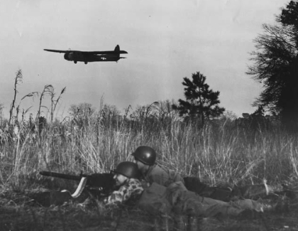 Soldiers crouch in high grass while a plane flies overhead.