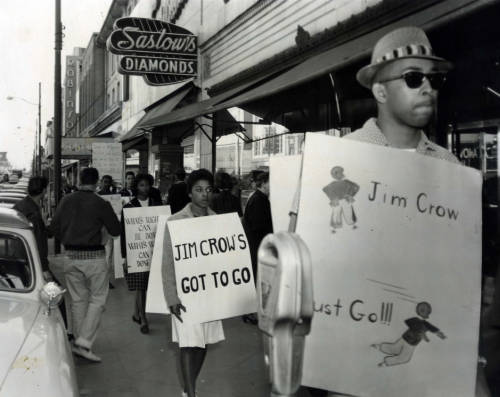 Male and female black protestors wearing signs, walking down a sidewalk under a &quot;Saslow's diamonds&quot; business sign. The protestors signs read: &quot;Jim Crow Must Go!&quot; &quot;Jim Crow's Got to Go!&quot; and &quot;What's right can be done ...&quot; Black and white photograph. 