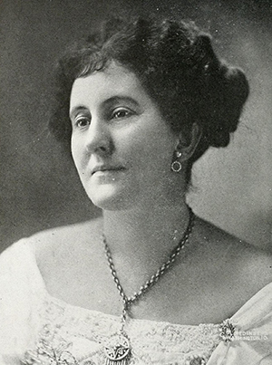 Katherine Hawkins. She is wearing a large necklace and dress. 