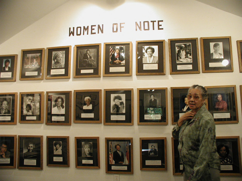 A woman stands in front of photographs of black women below a Women of Note sign