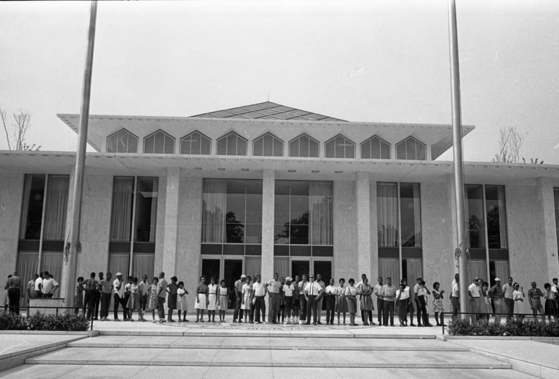 A group of Black people stand outside a large concrete building with tall windows
