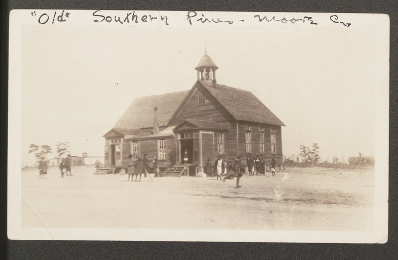 Black children around the outside of a dark wood building with a bell on the roof