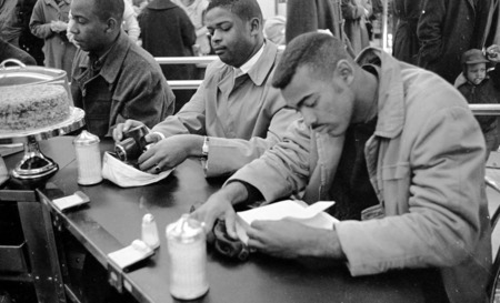 Three black men sit quietly at a lunch counter, one is reading, another has a camera