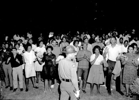 A police officer stands in front of a group of African Americans singing and clapping on a lawn