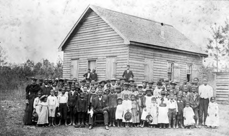 A large group of Black children and several adults stand in front of a wooden school building