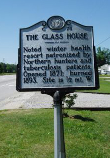 Large metallic marker that reads: &quot;THE GLASS HOUSE, Noted winter health resort patronized by Northern hunters and tuberculosis patients. Opened 1871; burned 1893. Site is 1/2 mi. W.&quot;