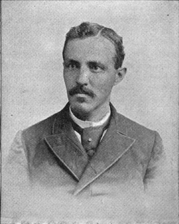Black and white photograph of John Crosby. He has a mustache and small chin beard, his hair parted on the right side. He is gazing to the left with a serious expression. He is wearing a fastened jacket, white shirt and tie.