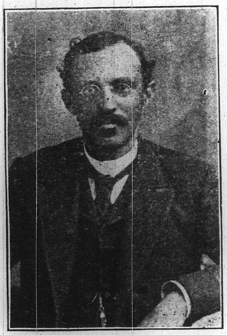 Crosby sits facing the camera. He has a mustache and soul patch. His hair is dark and parted on the right side. He is wearing round wire-rimmed glasses. He is dressed in a suit coat, vest, white button-up shirt and a tie.
