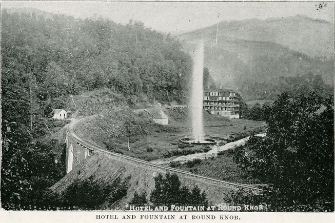 A large geyser erupts. Flanked to its left is a railway which runs along a viaduct. In the background a hotel can be seen. Wooded, mountainous terrain sets the photo.