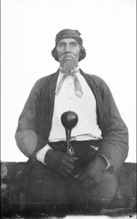 Swimmer is seated, holding a gourd rattle. He is wearing a button shirt, jacket, and pants with scarves around his head and neck. 